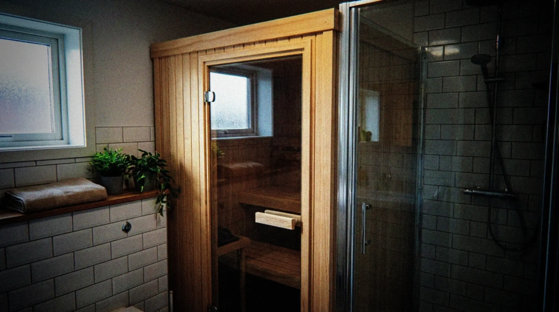 A compact wooden sauna installed in the corner of a small bathroom, showing how it fits neatly between a shower stall and wall. The sauna has a glass door revealing cedar interior with a single bench. Natural lighting comes through a small bathroom window, highlighting the warm wood tones contrasting with modern white bathroom tiles. A folded towel sits on a nearby shelf, with a few plants adding a touch of greenness to the space. The perspective shows the clever use of limited space in an ordinary apartment setting.
