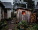 A small wooden outdoor sauna with steam rising from its chimney, situated in a backyard garden next to a cottage on an overcast afternoon. An electricity meter is visible on the side of the cottage, and a small pile of firewood is stacked neatly nearby. Through the sauna's small window, a warm glow emanates, creating contrast with the cooler outdoor tones. The sauna appears well-maintained but not brand new, suggesting regular use. A simple garden path connects the cottage to the sauna structure, with some potted plants and natural landscaping surrounding the area.