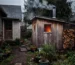 A small wooden outdoor sauna with steam rising from its chimney, situated in a backyard garden next to a cottage on an overcast afternoon. An electricity meter is visible on the side of the cottage, and a small pile of firewood is stacked neatly nearby. Through the sauna's small window, a warm glow emanates, creating contrast with the cooler outdoor tones. The sauna appears well-maintained but not brand new, suggesting regular use. A simple garden path connects the cottage to the sauna structure, with some potted plants and natural landscaping surrounding the area.